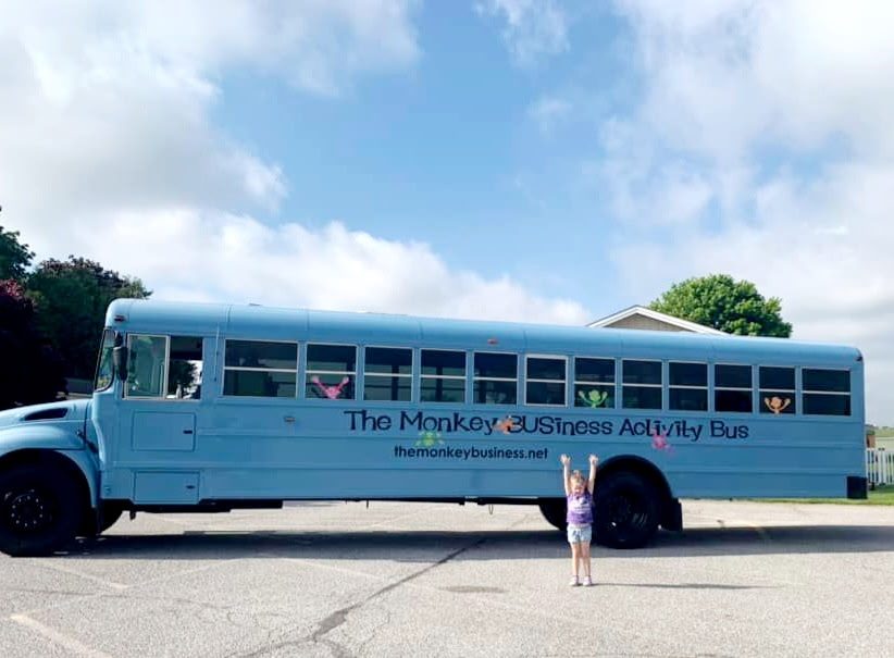 The Monkey BUSiness Activity Bus, a light blue converted school bus, parked with a child waving her arms in front
